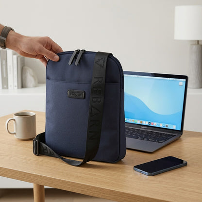 Navy Bugatti Reborn Slim Crossbody Bag on a desk beside a laptop and phone for scale reference.