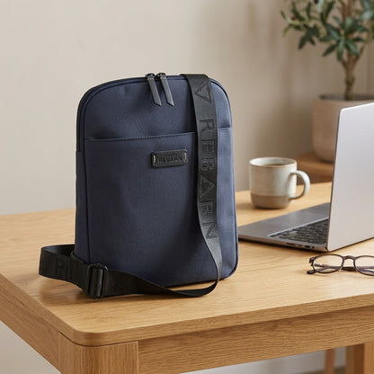 Bugatti Reborn navy slim crossbody bag on a wooden desk with a laptop, coffee mug, and glasses.
