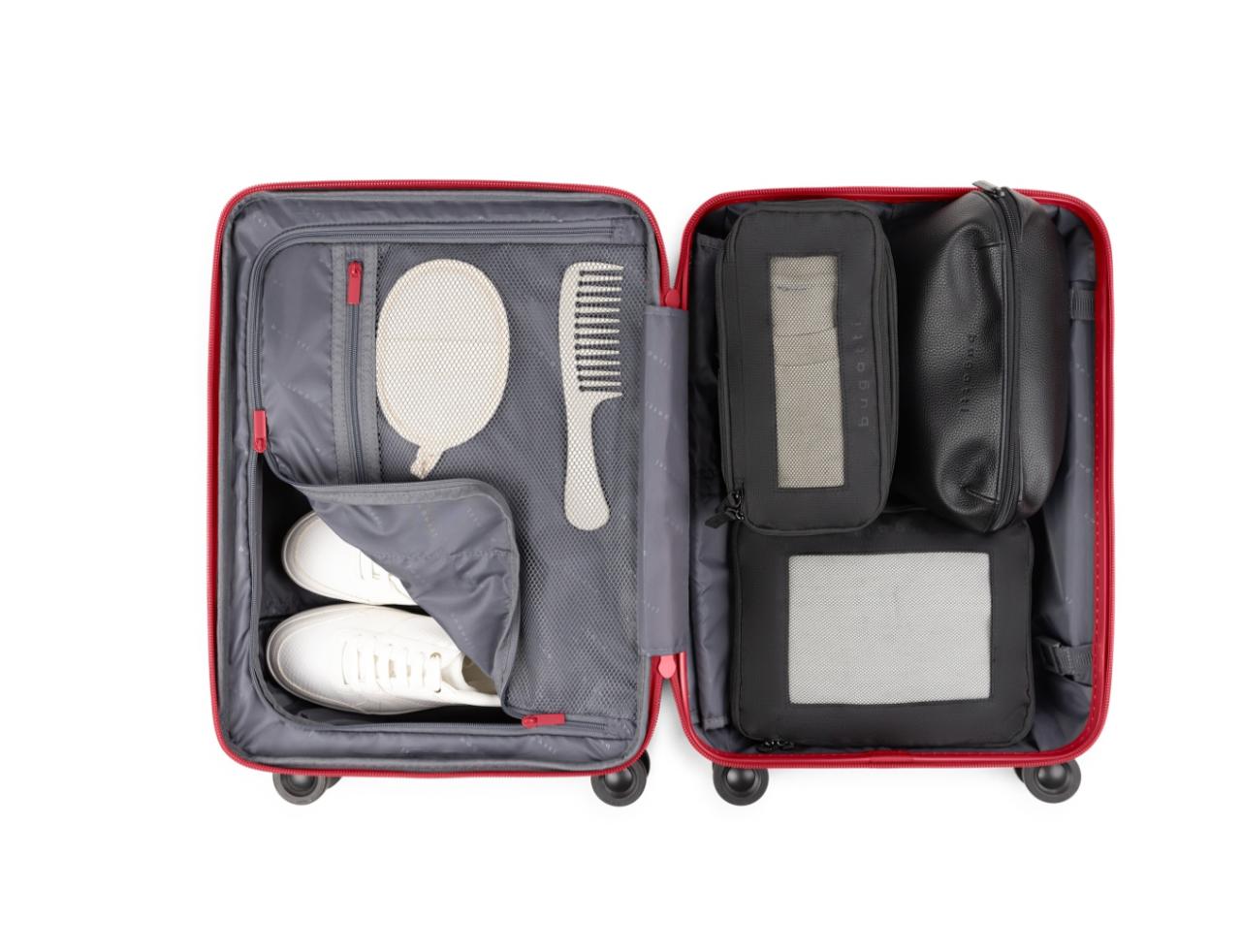 An open red hard-shell suitcase shown from above, packed with gray toiletry organizers, a white hand mirror, a white comb, and a pair of white shoes on a plain white background.